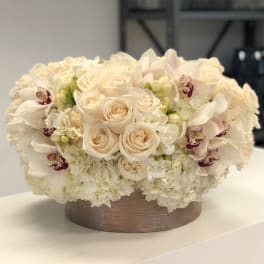 Low round arrangement of cream roses, hydrangeas, and orchids in a short metallic container