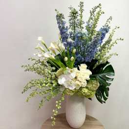 Tall arrangement of white roses, orchids, calla lilies and blue delphinium in a white vase