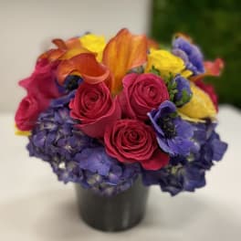 Low round arrangement of hot pink roses, orange calla lilies, yellow roses and purple blooms in a black vase