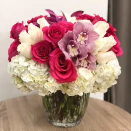 Round arrangement of hot pink roses, white roses, orchids, and hydrangeas in a clear glass vase.