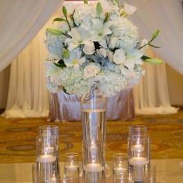 White floral centerpiece in a tall glass vase with candles around it