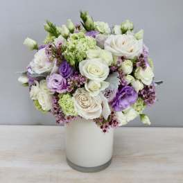 Round arrangement of white roses and lavender blooms in a white cylinder vase