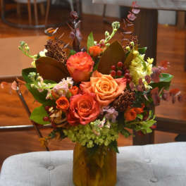Autumn-toned bouquet of orange roses and mixed blooms in an amber glass jar vase