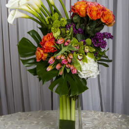Tall bouquet of white calla lilies, orange roses, and purple flowers in a glass vase