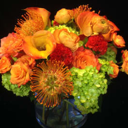 Low round arrangement of orange calla lilies, roses, red blooms and green hydrangeas in a clear glass vase
