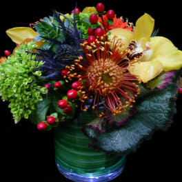 Low arrangement of green hydrangea, orange protea, yellow orchid, succulents, and red berries in a glass vase