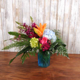 Colorful mixed arrangement with lilies, hydrangeas, and a rose in a blue glass vase on a wooden surface