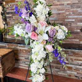 Cross-shaped floral tribute with white lilies, roses, and purple flowers on an easel