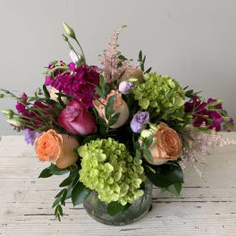 Low mixed arrangement of peach and pink roses, green hydrangeas, and purple blooms in a clear round vase