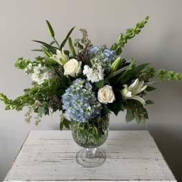 Large white and blue flower arrangement with lilies, roses and hydrangeas in a clear glass pedestal vase