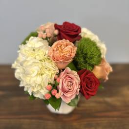 Short round arrangement of red and pink roses, peach blooms, white hydrangea and green dianthus in a white vase