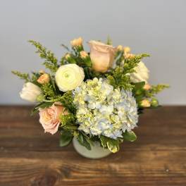 Low arrangement of blue hydrangea, white ranunculus, and peach roses in a round white vase.