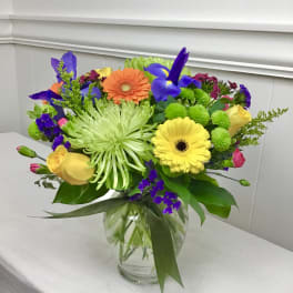 Bright mixed bouquet with yellow roses, gerbera daisies, green spider mums, and purple irises in a glass vase.