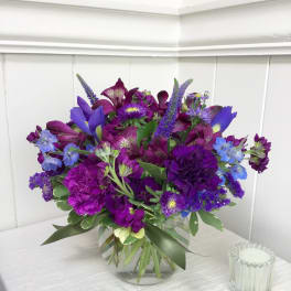 Round arrangement of purple and blue flowers in a clear glass vase on a white table.