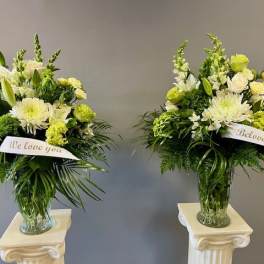 Two white and green sympathy flower arrangements in glass vases with ribbon banners