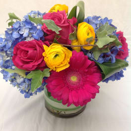 Bright arrangement of pink roses and gerbera daisy with yellow blooms and blue hydrangeas in a glass vase