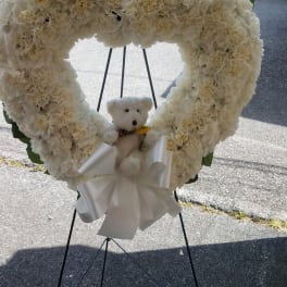 Heart-shaped white floral wreath with a teddy bear and ribbon on a stand