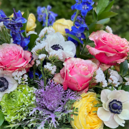 Colorful arrangement with pink roses, white anemones, blue delphinium, and other mixed blooms.
