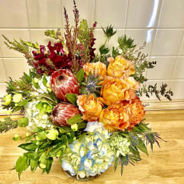 Mixed bouquet with orange roses, blue hydrangea, and burgundy protea in a low vase