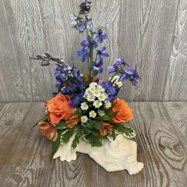 Orange roses, blue delphinium, and white daisies in a white ceramic turtle container