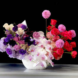 Asymmetrical bowl arrangement of pink, purple and red roses, dahlias and orchids in a white vessel.