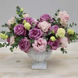 Low centerpiece of purple roses with pale pink and green carnations in a white footed bowl.