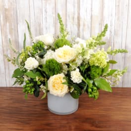 Low vase arrangement of cream roses, white flowers, and green accents in a frosted cylinder container.