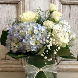Blue hydrangeas and pale yellow roses in a green glass vase with a ribbon