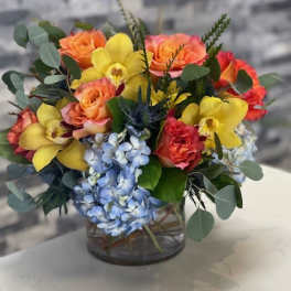 Short arrangement of orange roses, yellow orchids, and blue hydrangeas in a clear glass vase.