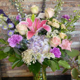 Mixed pastel bouquet with lilies, roses, hydrangea, and purple accents in a glass vase