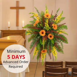 Tall sympathy easel spray with orange gerbera daisies, yellow blooms, and ferns beside a casket.