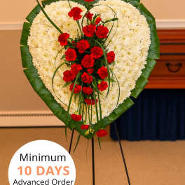 Heart-shaped standing wreath of white flowers with red carnations and a green leaf border on an easel.