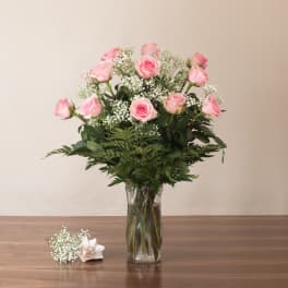 Tall glass vase of light pink roses with white filler flowers and a small matching corsage on the table.