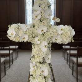 Tall white floral cross with orchids and roses displayed on an easel in a chapel