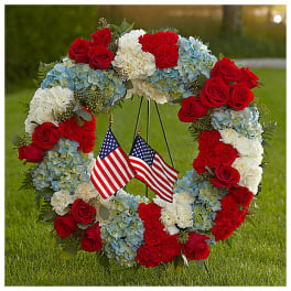 Floral wreath with red roses, blue hydrangeas, and white carnations around two small American flags