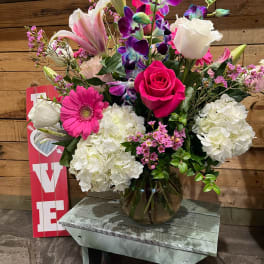 Mixed arrangement of pink, white, and purple flowers in a glass bowl vase on a rustic wooden bench
