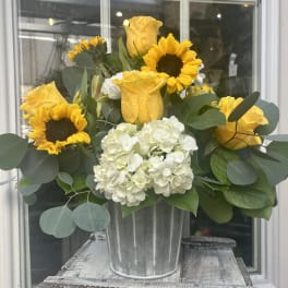 Yellow roses, sunflowers, and white hydrangeas arranged in a metal bucket-style vase