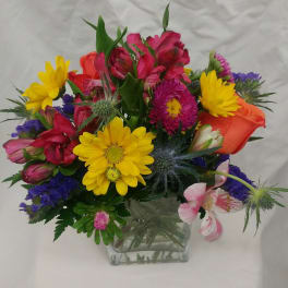 Mixed bouquet in a clear square vase with yellow daisies and pink, orange, and purple blooms