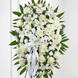 Tall standing spray of white lilies, roses, and carnations with white ribbon on a green leaf backdrop