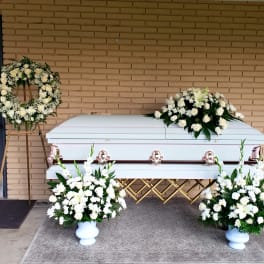 White casket with white floral sprays and a wreath on easels