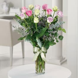 Tall glass vase of pink, white, and lavender roses with purple filler flowers and a ribbon bow.