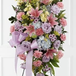 Standing spray of pink lilies, carnations, lavender roses, and blue hydrangeas with lavender ribbons on an easel.