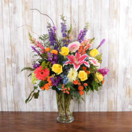 Tall mixed arrangement with yellow roses, pink lilies, gerbera daisies and purple flowers in a clear glass vase