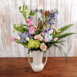 Tall mixed flower arrangement in a white ceramic pitcher with pink, blue, and green blooms