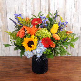 Bright mixed arrangement with sunflower, lilies, roses and gerbera daisies in a dark glass vase