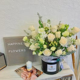Cream roses and white blooms arranged in a black hat box with a ribbon.