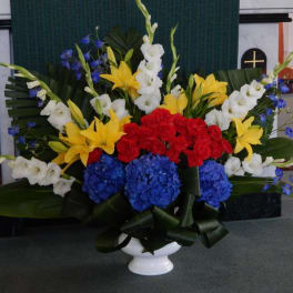 Large funeral arrangement with red, yellow, white, and blue flowers in a white vase