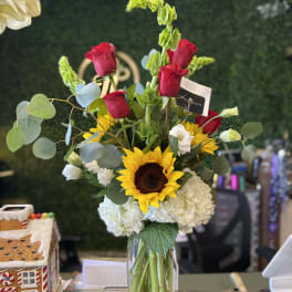 Mixed bouquet in a clear glass vase with red roses, a sunflower, and white hydrangeas.
