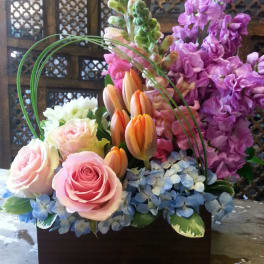 Mixed bouquet of pink roses, orange tulips, and purple blooms in a dark container