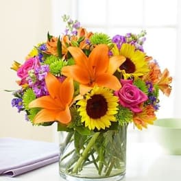 Mixed bouquet of orange lilies, sunflowers, and pink roses in a glass vase
